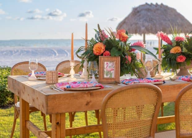 A wooden outdoor dining table is set with colorful floral arrangements, glassware, and napkins, overlooking a beach with a thatched umbrella in the background.