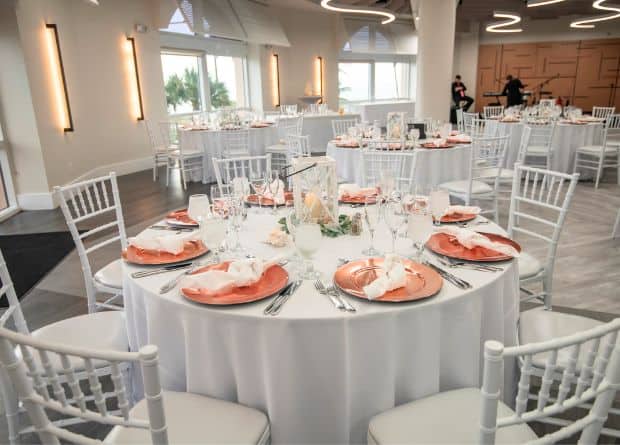 Round tables with white tablecloths and white chairs are set for an event, with peach plates, folded napkins, and glassware in a bright, modern room.