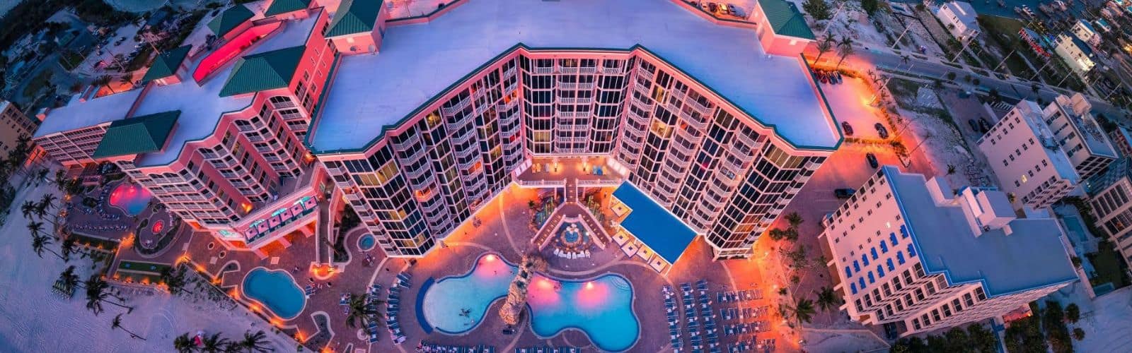 Aerial view of a large beachfront resort at dusk, featuring multiple pools, palm trees, and brightly lit buildings with balconies facing the ocean.