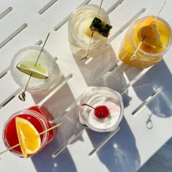 Five cocktails in clear glasses, each garnished with fruit or a cherry, arranged on a white slatted surface in sunlight.