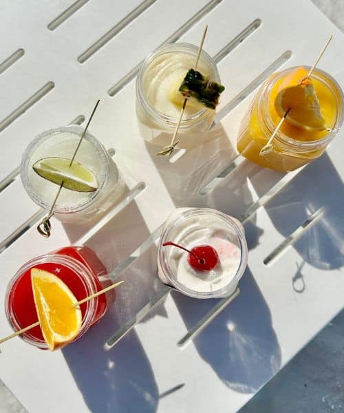 Five cocktails in clear glasses, each garnished with fruit or a cherry, arranged on a white slatted surface in sunlight.