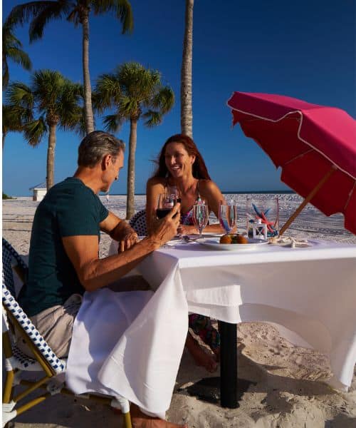 A man and woman sit at a table covered with a white cloth, dining and drinking wine on a sandy beach with palm trees and a red umbrella under a clear blue sky.