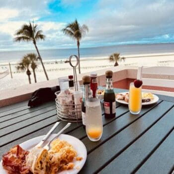 Breakfast plates and drinks on a table overlooking a sandy beach with palm trees and a cloudy blue sky in the background.
