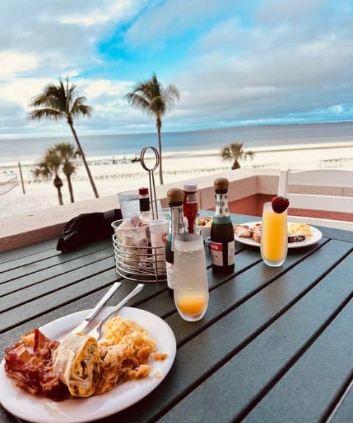 Breakfast plates and drinks on a table overlooking a sandy beach with palm trees and a cloudy blue sky in the background.