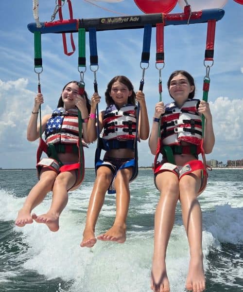 Three girls wearing life jackets are parasailing together over the ocean, with buildings and clouds visible in the background.