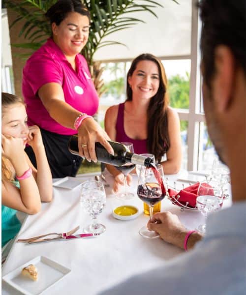 A server pours red wine into a glass at a restaurant table where three people sit, with glasses of water and plates in front of them.