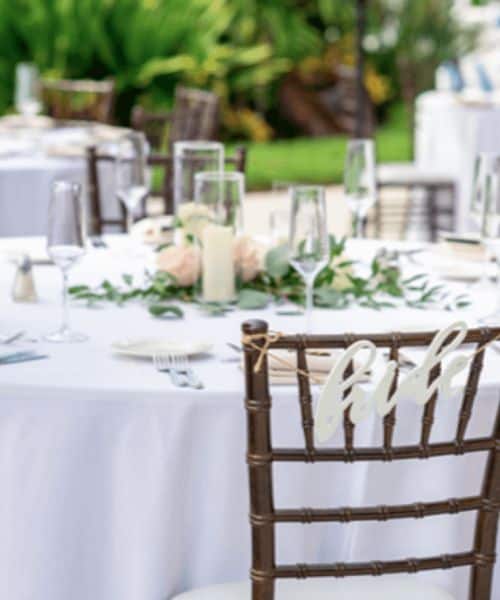 A round table set for an outdoor event with white tablecloth, glassware, plates, a floral centerpiece, and a chair labeled “bride.”.