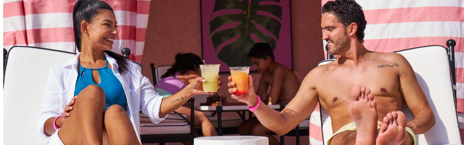 A woman and man relax on lounge chairs by a pool, smiling and clinking drinks, while two people play chess in the background.
