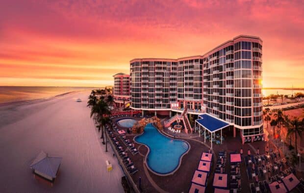A beachfront resort with a curved multi-story building, pool area, palm trees, and lounge chairs, overlooks a sandy beach at sunset.