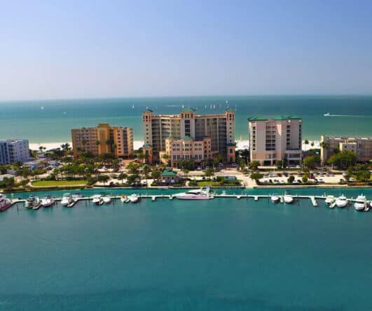 Aerial view of waterfront hotels, boats docked at a marina, and a sandy beach with the ocean in the background under a clear sky.