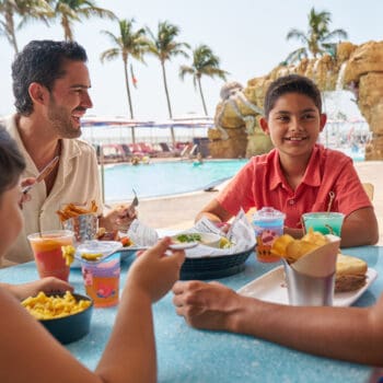 A family of four eats lunch together at an outdoor table near a pool, with palm trees and lounge chairs in the background.