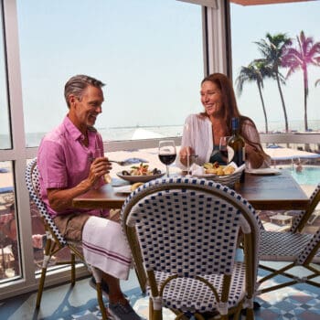A man and a woman sit at a restaurant table by a window, eating food and drinking wine, with a pool and palm trees visible outside.