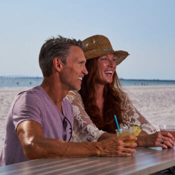 A man and woman sit at a beachside bar, smiling and holding drinks. The woman wears a straw hat and sunglasses are on the table. The beach and ocean are in the background.