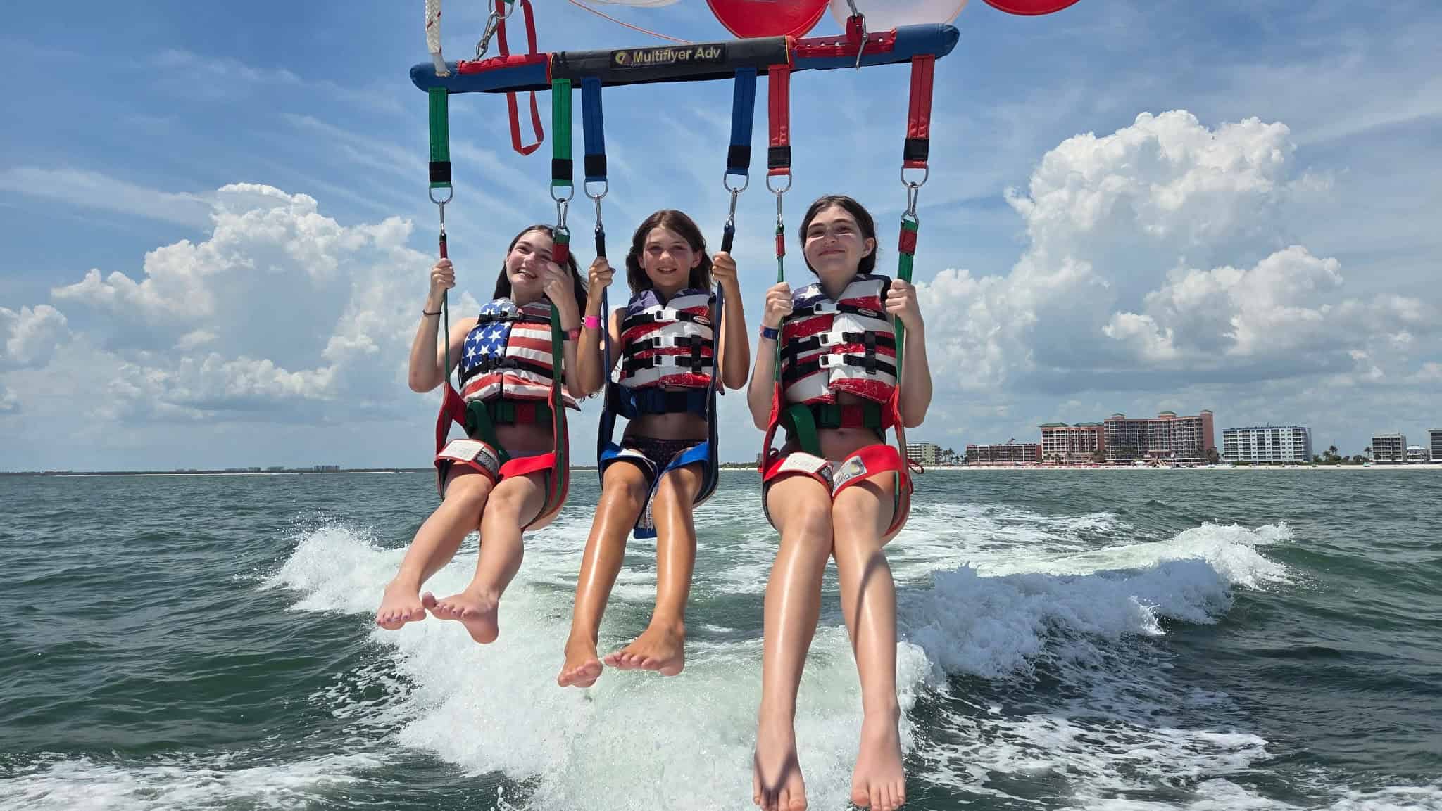 Three girls wearing life jackets and harnesses are parasailing above the ocean, with buildings and clouds visible in the background.