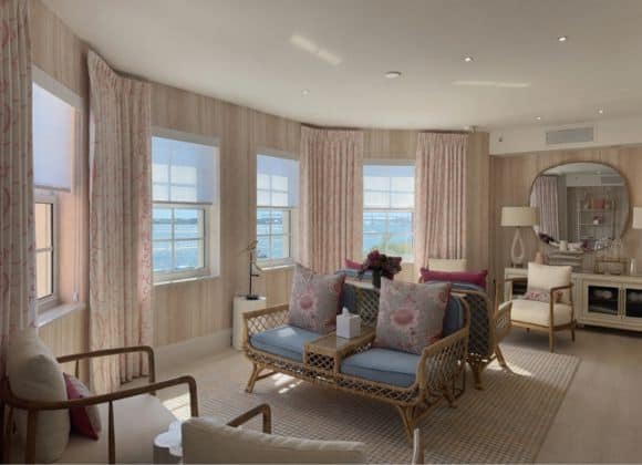 Bright living room with large windows, light wood flooring, wicker furniture with pastel cushions, patterned curtains, and a view of the ocean outside.