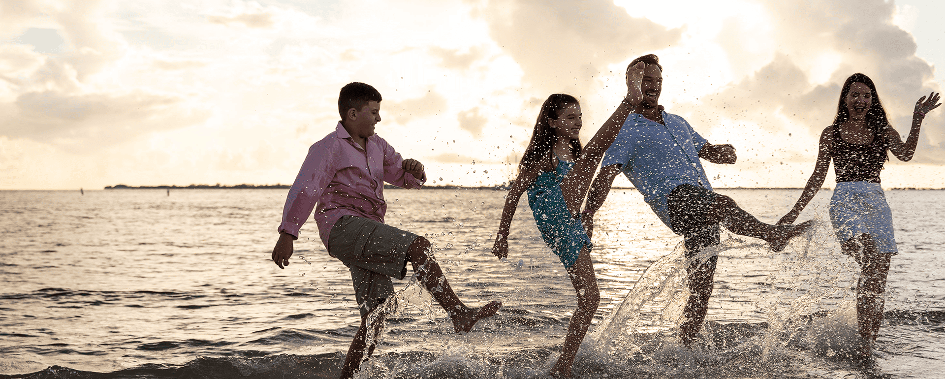 Four people stand in shallow water at the beach, kicking up splashes and smiling, with the sun low in the sky behind them.
