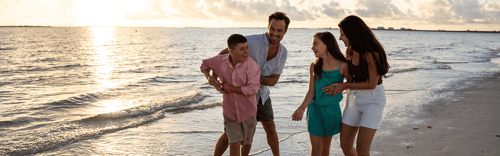 Four people, two adults and two children, walk and smile together on a sandy beach near the water during sunset.
