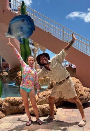 A girl and a man pose with arms outstretched near a pool under a blue sky, standing by a large decorative fish and tropical-themed structures.