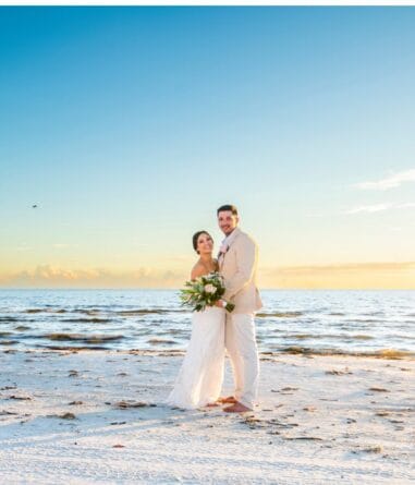 A bride and groom stand together on a sandy beach at sunset, holding a bouquet, with the ocean and blue sky in the background.