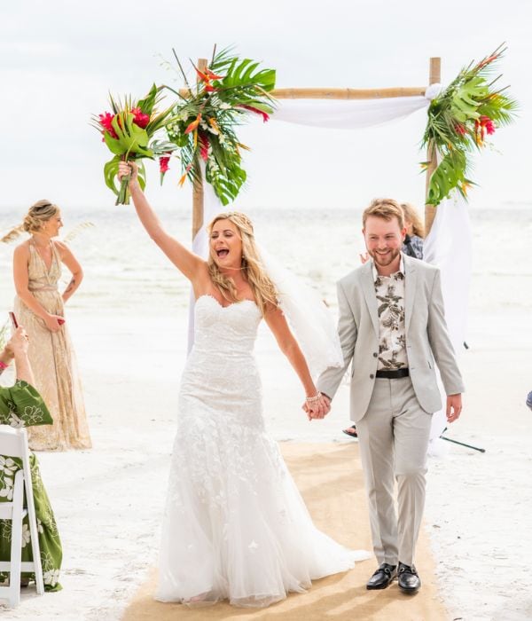A bride and groom walk down a sandy aisle at a beach wedding, with the bride raising her bouquet and the groom smiling, under a decorated bamboo arch.
