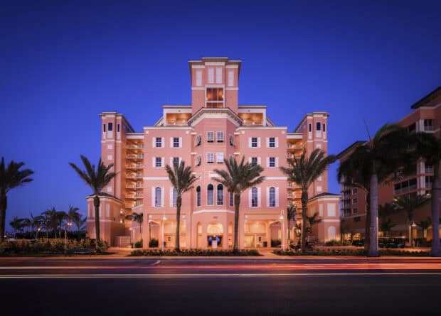 Pink multi-story hotel with palm trees in front, illuminated at dusk with streaks of car lights passing by on the street.