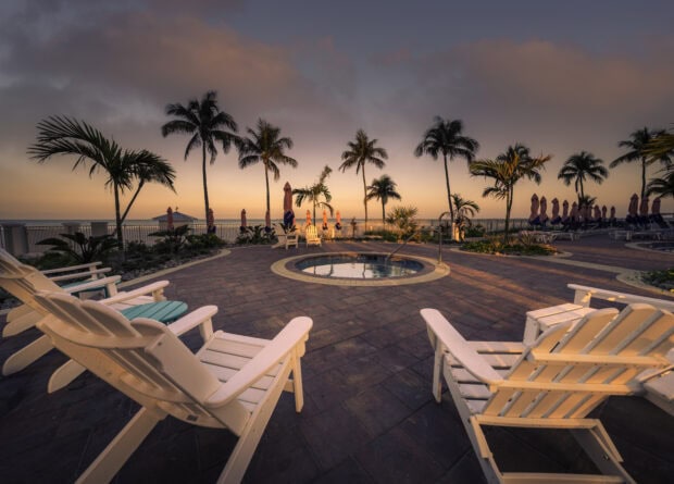 Lounge chairs posed around a hot tub, with palm trees and the beach in the distance - at dusk, the sky casting a pink glow over the scene