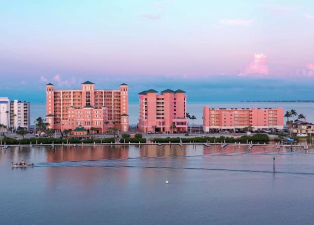 Three pink mid-rise buildings stand along the waterfront, with a boat passing by in the foreground and a calm bay and sky in the background.