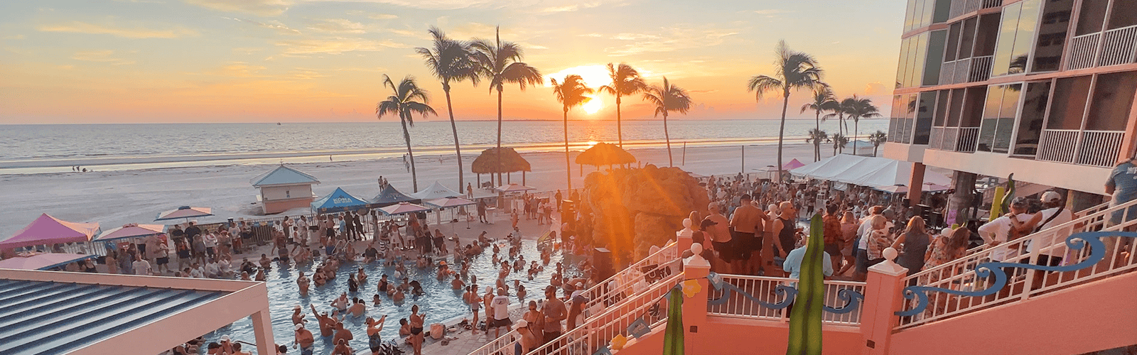 Crowd gathered around a beachside pool and on terraces at sunset, with palm trees, tents, and the ocean visible in the background.