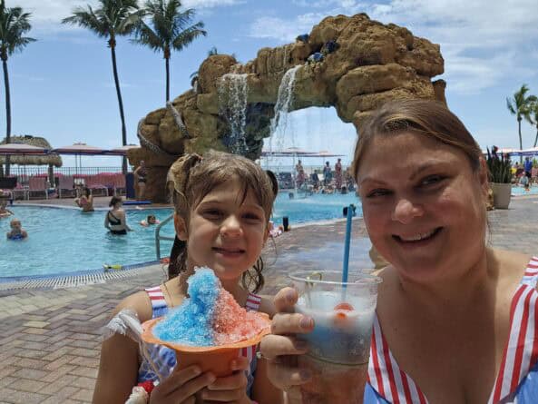 A woman and a girl smile by a pool, holding a red, white, and blue shaved ice and a cup with a drink; a rock waterfall and palm trees are in the background.