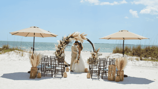A bride and groom kiss under a floral arch at a beach wedding setup with chairs, pampas grass arrangements, and umbrellas on the sand by the ocean.