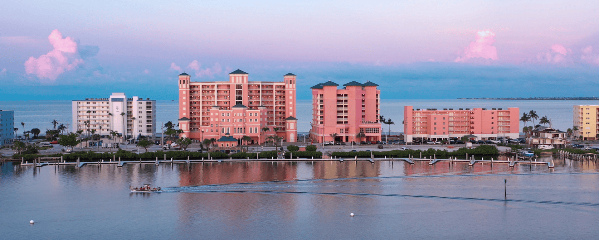 Waterfront view of pastel-colored hotels and condos along a calm bay at sunset, with a small boat creating a wake in the water.