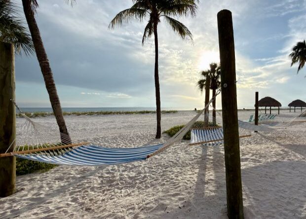Empty striped hammocks hang between wooden posts on a sandy beach with palm trees, overlooking the ocean under a partly cloudy sky.