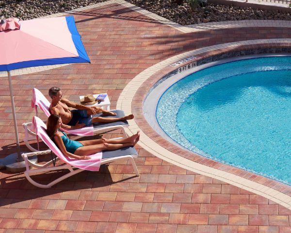 Two people recline on lounge chairs under a pink umbrella beside a curved outdoor swimming pool with clear blue water.