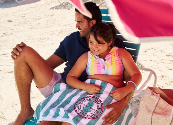 A man and a smiling girl sit on a beach chair at the beach, with a striped "Pink Shell Beach Club" towel covering their laps.