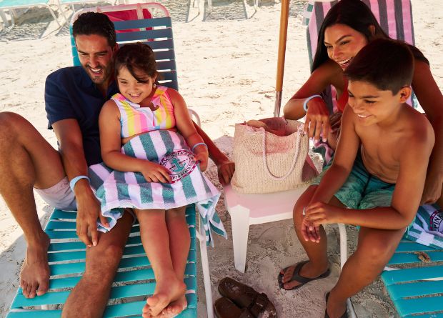 A family of four sits on lounge chairs at the beach, smiling and relaxing together with towels and beach accessories.