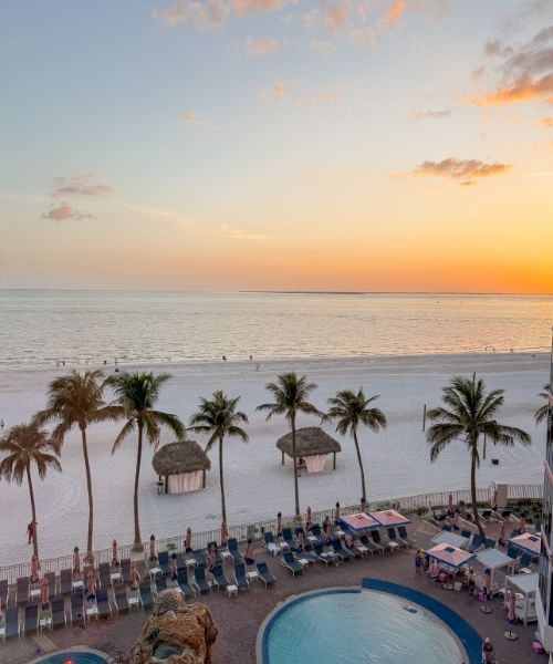 Palm trees, lounge chairs, and cabanas line a sandy beach beside a pool, with a calm ocean and colorful sunset sky in the background.