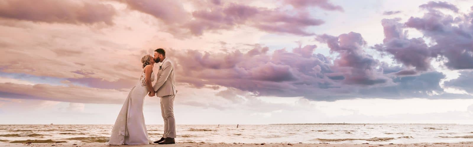 A bride and groom stand on a beach facing each other under a dramatic cloudy sky at sunset.