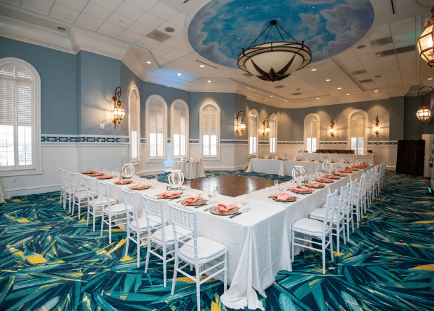 A banquet room with a U-shaped table arrangement, white chairs, pink napkins, and a decorative blue-green carpet under a ceiling mural and large chandeliers.