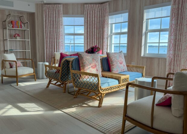 Bright sitting area with wicker chairs, cushioned seats, and large windows overlooking water; light wooden floors and pink-accented decor complete the room.