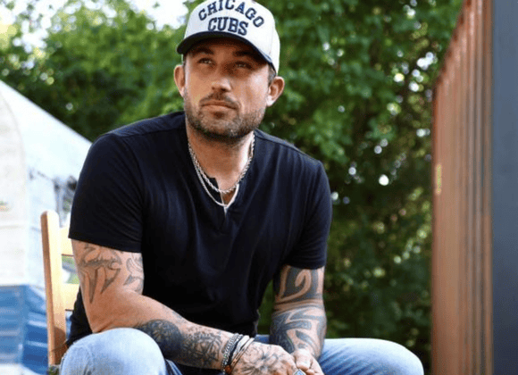A man with tattoos sits outdoors on a chair, wearing a black t-shirt, a Chicago Cubs hat, jeans, necklaces, and bracelets, with trees and a building in the background.