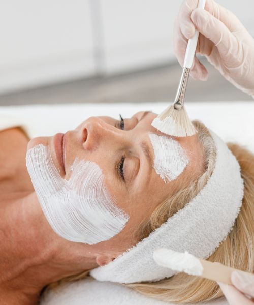 A woman with a headband receives a facial treatment as a white cream is applied to her face with brushes by a gloved person.