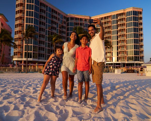 A family of four stands on a sandy beach in front of a tall hotel, posing for a selfie in warm sunlight.