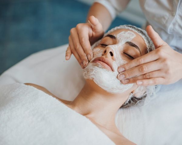 Person lying on a spa bed receives a facial treatment as an esthetician applies a white facial mask to their face using their hands.