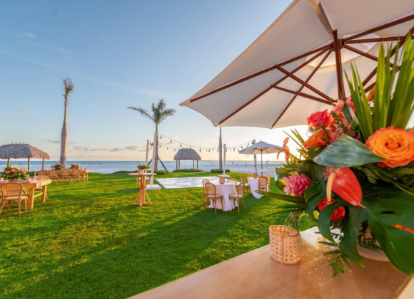 Outdoor beachside event setup with tables, chairs, large umbrellas, tropical flowers, palm trees, and the ocean in the background at sunset.