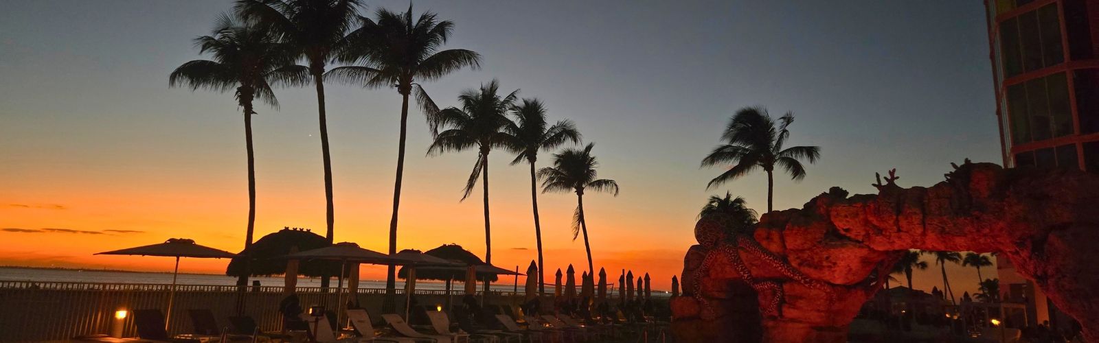 Palm trees and poolside lounge chairs are silhouetted against an orange sunset sky, with a rock formation and umbrellas in the foreground.