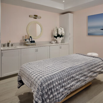 A spa treatment room featuring a massage table covered with a textured grey blanket, a vanity with a sink and round mirror, and framed wall art of stacked stones.