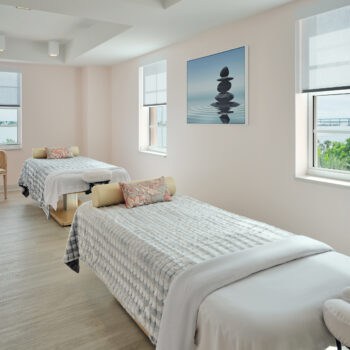 Two massage tables covered in blankets and bolsters in a bright room with windows overlooking a bay.