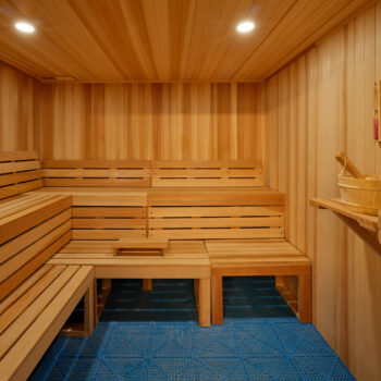 A wood-paneled sauna featuring tiered benches and a blue-gridded floor, with a wooden bucket and ladle on a side shelf.