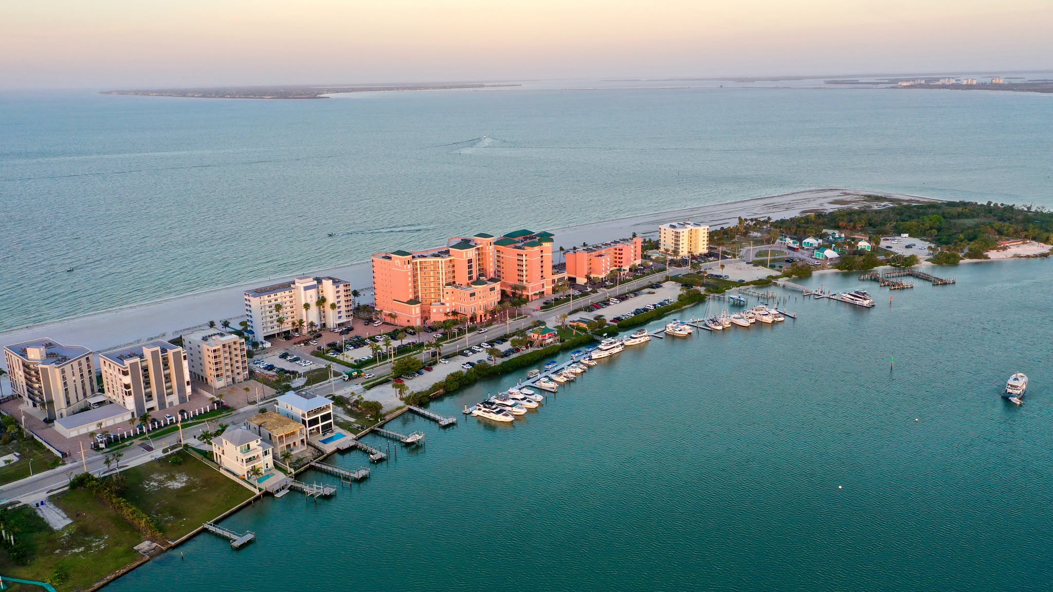 Aerial view of a coastal resort area with multi-story apartment buildings, a marina filled with boats, and a sandy beach along the water.
