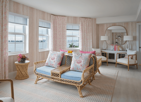 A living area featuring two rattan-framed loveseats with blue cushions and floral pillows, facing a console table with a large round mirror against a wall with pink patterned wallpaper and curtains.